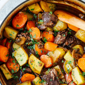 A white Dutch oven filled with hearty Dutch Oven Beef Stew, featuring chunks of beef, potatoes, carrots, and herbs in a rich brown broth. A wooden spoon rests in the pot.
