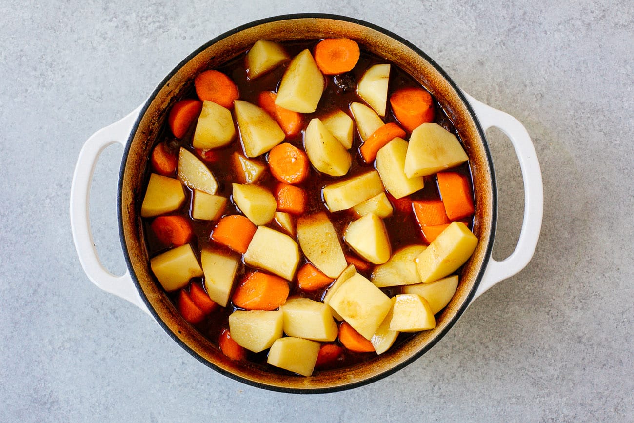 A white pot filled with chopped potatoes and carrots in a savory brown broth, perfect for making Dutch Oven Beef Stew, set on a light gray surface.