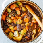 A white pot filled with Dutch Oven Beef Stew showcases tender chunks of beef, carrots, potatoes, and herbs in a rich brown broth. A wooden spoon rests inside the pot, with a striped cloth nearby.