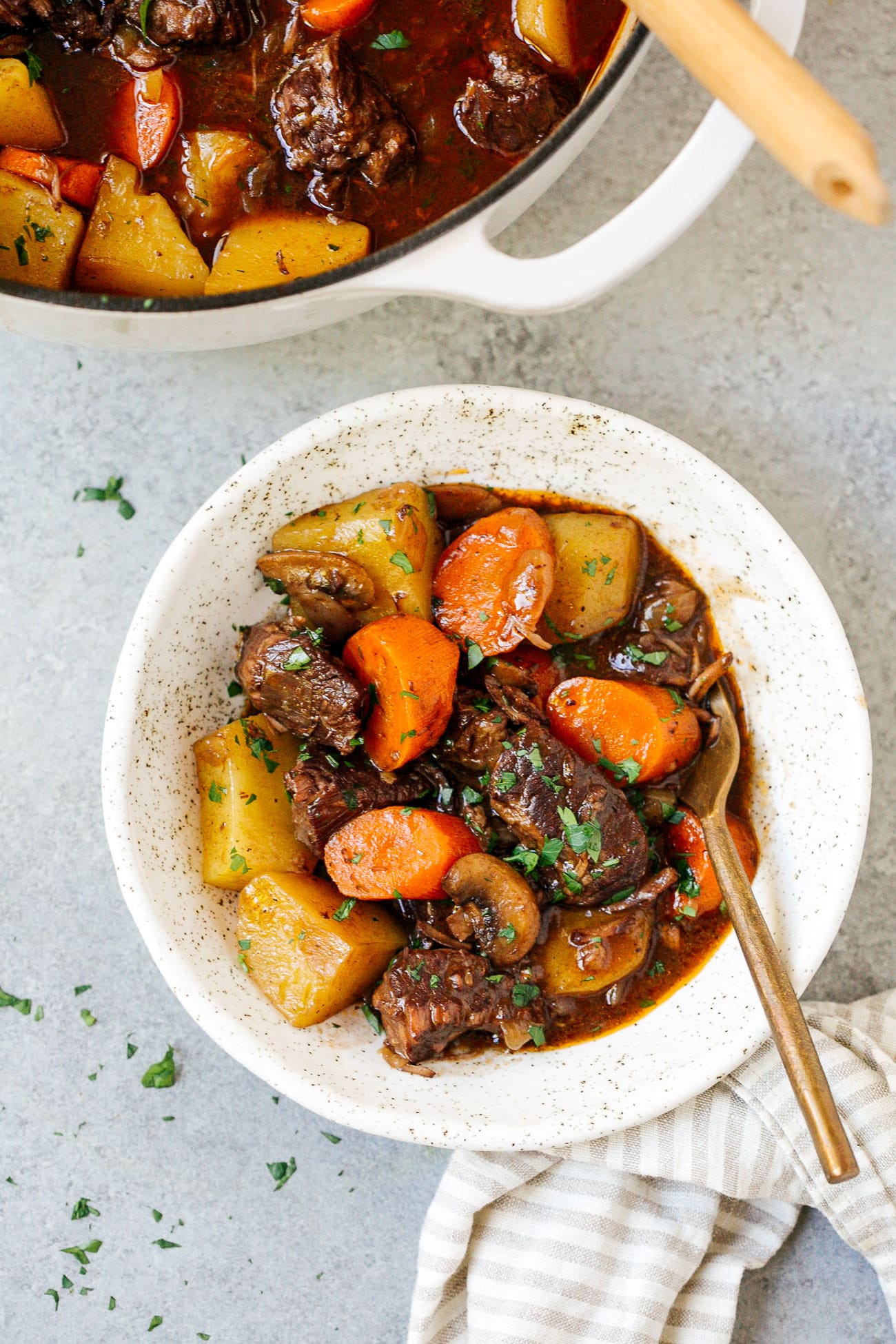 A bowl of Dutch Oven Beef Stew with chunks of beef, carrots, and potatoes in a rich brown broth, garnished with chopped parsley. A pot of stew and a striped napkin are nearby.