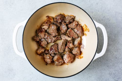 Chunks of browned beef are searing in a large, round, white Dutch oven on a light gray countertop, forming the rich base for a classic Dutch Oven Beef Stew with caramelized meat and savory juices collecting in the pot.