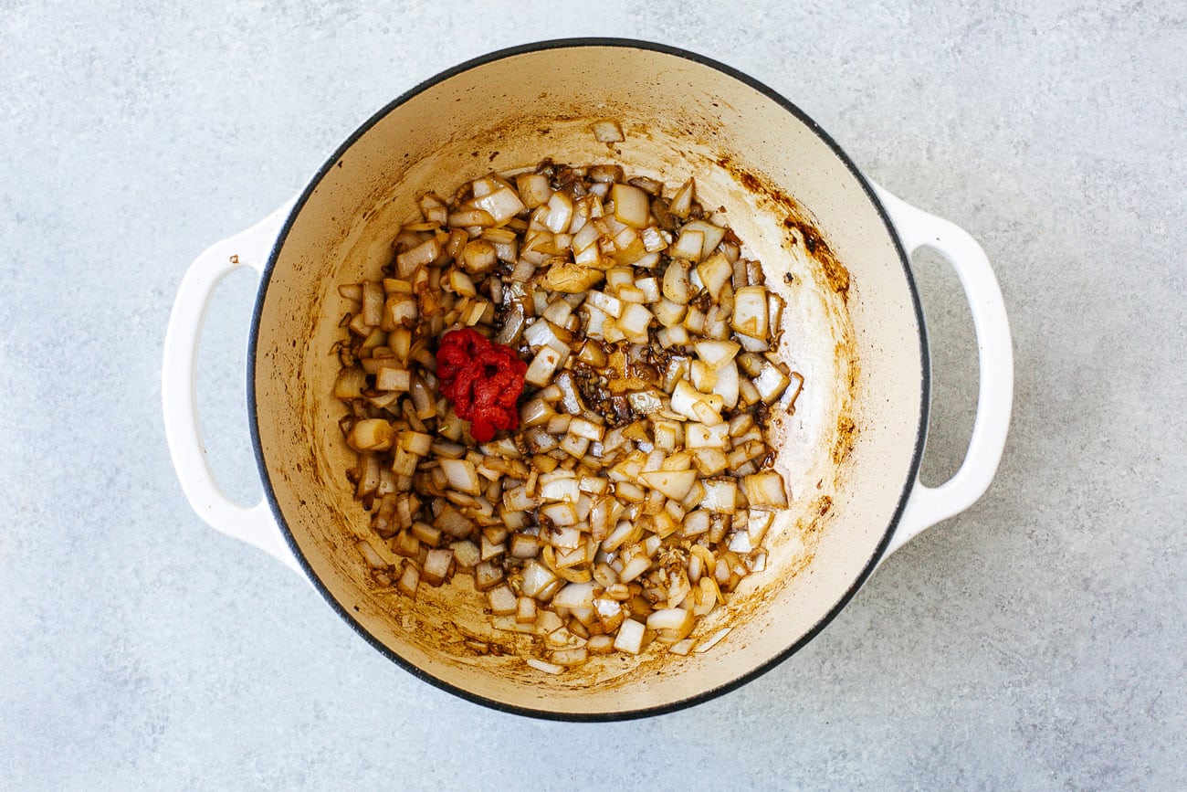 A white Dutch oven, perfect for Dutch Oven Beef Stew, contains sautéed chopped onions with a dollop of tomato paste in the center, resting on a light gray surface.