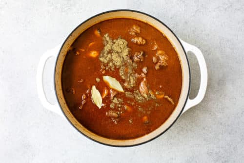 A white pot filled with Dutch Oven Beef Stew—rich brown broth, tender meat chunks, two bay leaves, and visible spices on top—set on a light gray surface.