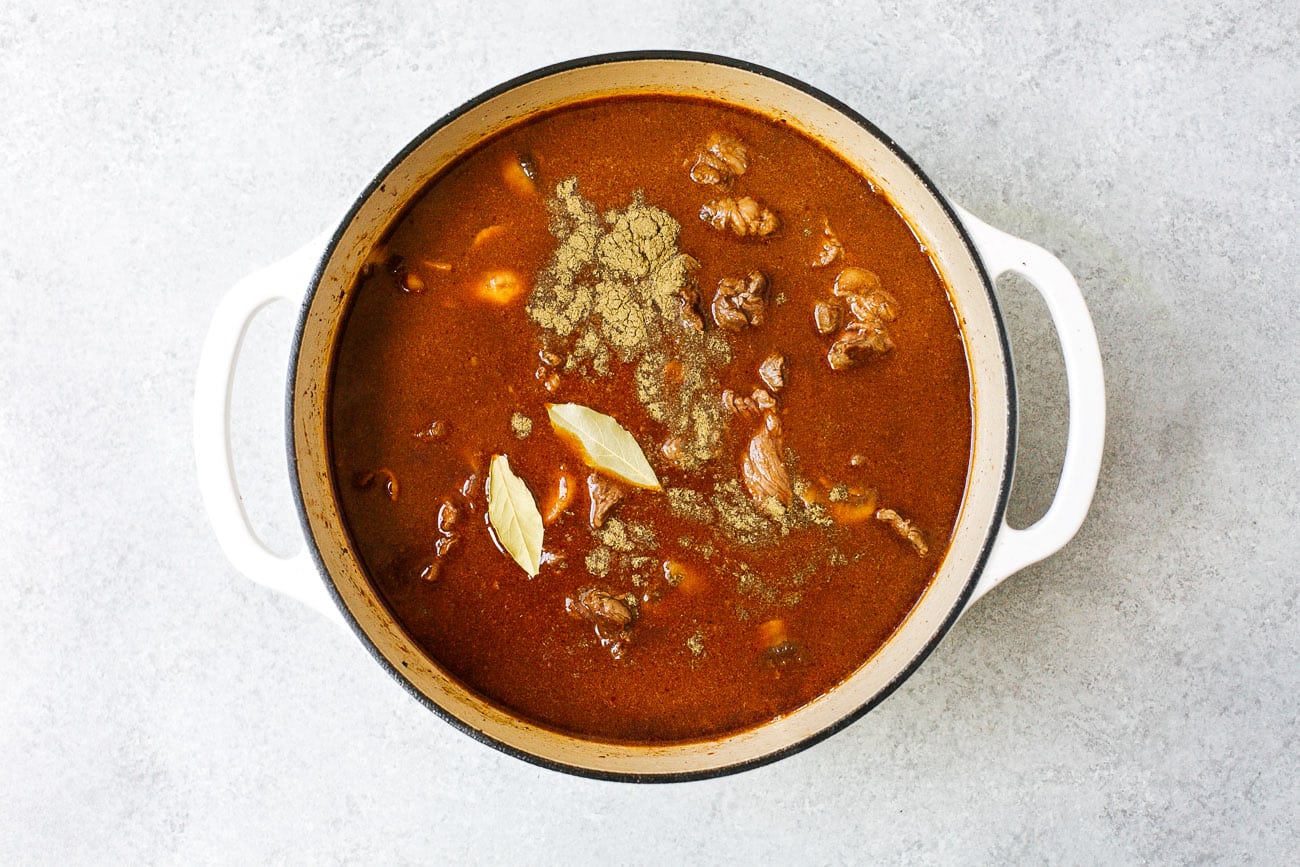 A white pot filled with Dutch Oven Beef Stew—rich brown broth, tender meat chunks, two bay leaves, and visible spices on top—set on a light gray surface.