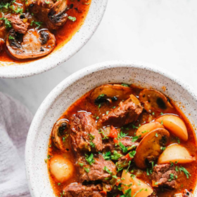 Two white bowls filled with Instant Pot Beef Stew, featuring tender chunks of beef, mushrooms, potatoes, and carrots in a rich red broth, garnished with chopped herbs. A striped napkin is beside the bowls.