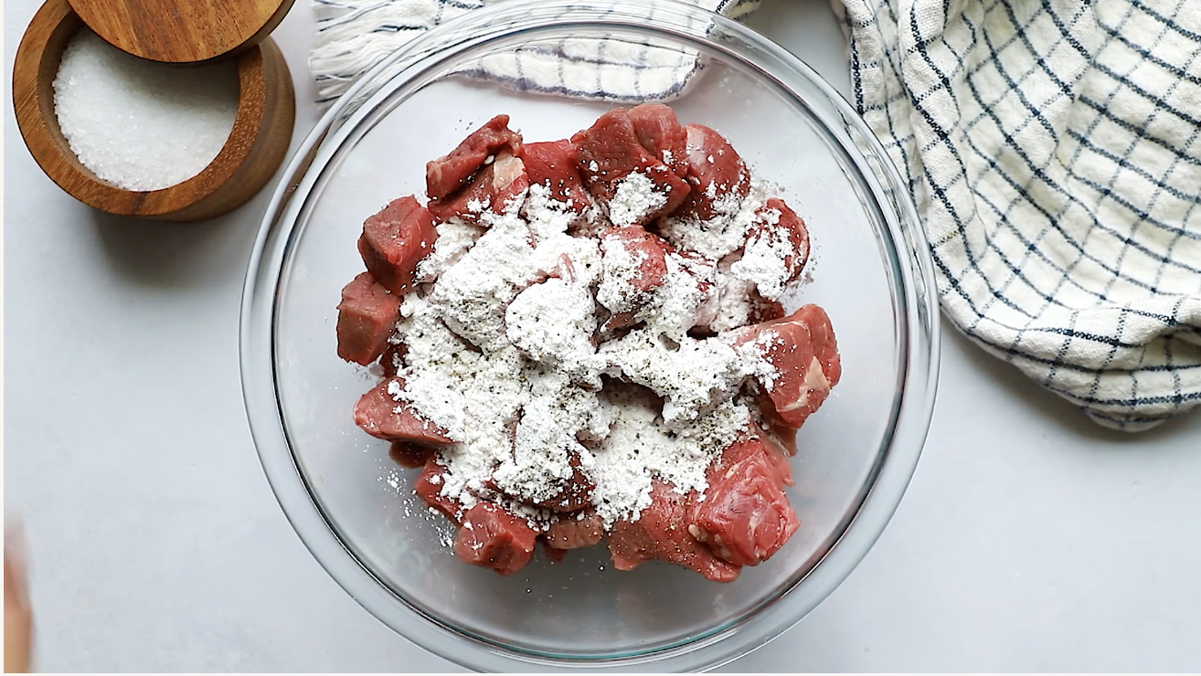 A glass bowl filled with raw beef cubes coated in flour, ready to be transformed into a hearty Instant Pot Beef Stew, sits next to a wooden salt container and a white kitchen towel with blue stripes on a light countertop.