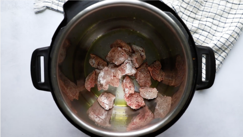 Chunks of raw beef with a light dusting of flour are in the bottom of a stainless steel pot, ready to be transformed into a hearty Instant Pot Beef Stew, next to a black-and-white checkered kitchen towel.