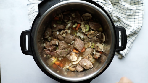 Top-down view of Instant Pot Beef Stew featuring chunks of beef, mushrooms, carrots, celery, and onions simmering inside an Instant Pot, next to a white and blue checkered dish towel.