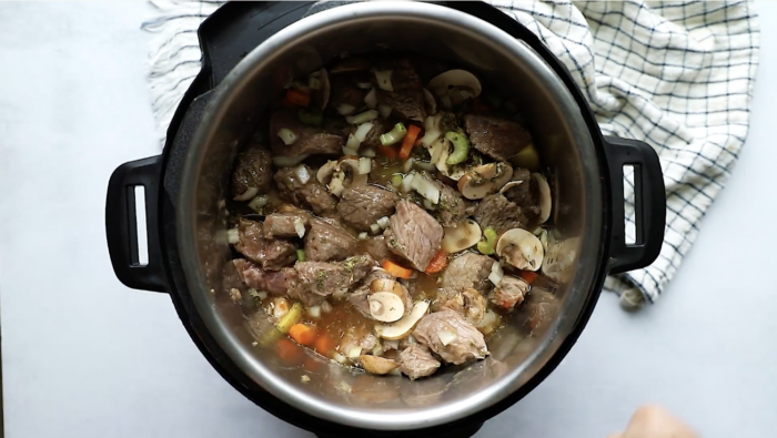 Top-down view of Instant Pot Beef Stew featuring chunks of beef, mushrooms, carrots, celery, and onions simmering inside an Instant Pot, next to a white and blue checkered dish towel.