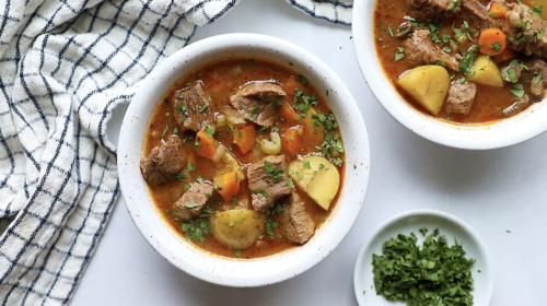 Two white bowls filled with hearty Instant Pot Beef Stew, brimming with tender chunks of beef, potatoes, and carrots, and garnished with chopped herbs. A small dish of herbs and a blue-and-white checkered cloth are nearby on a white surface.
