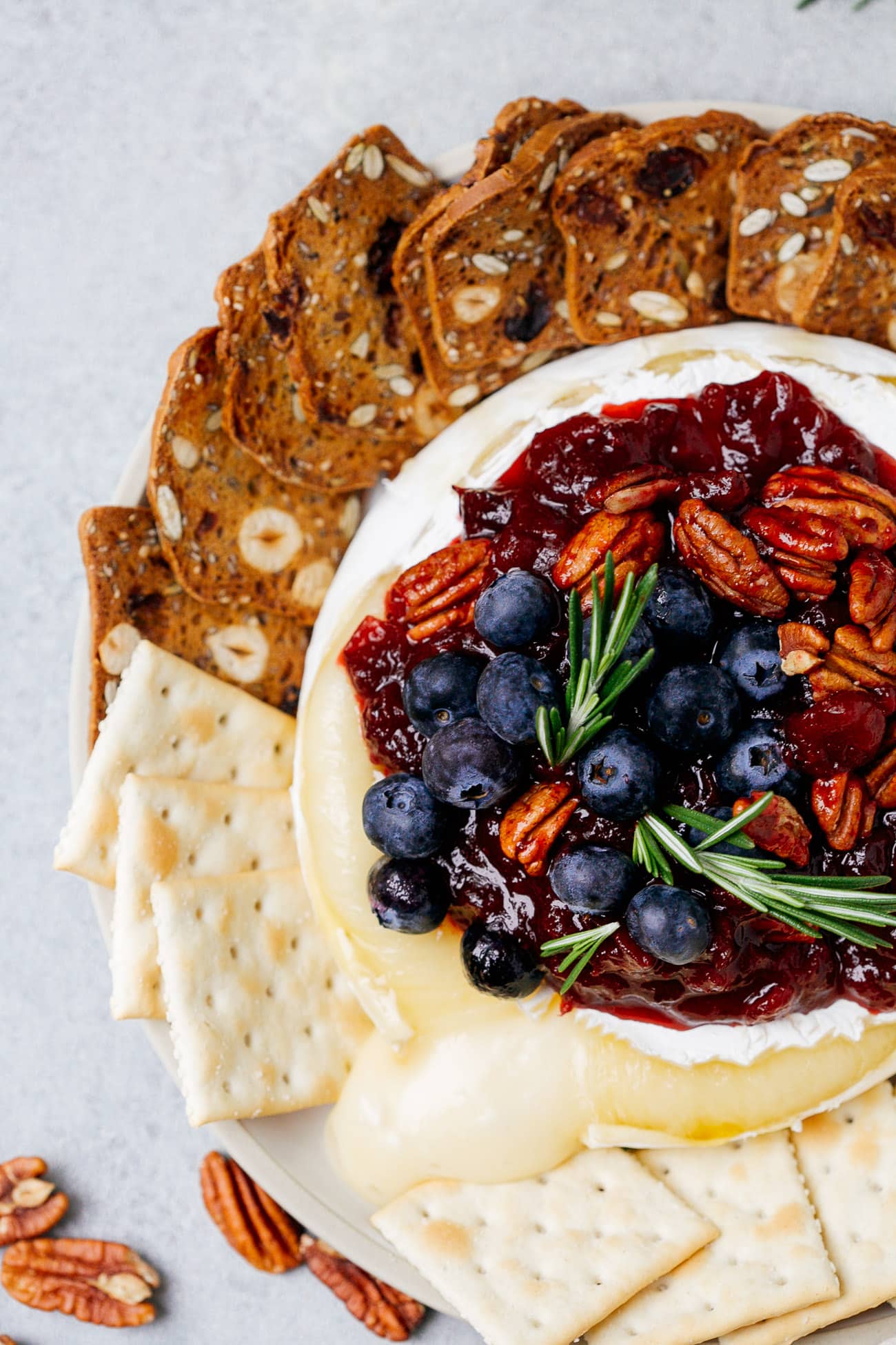 Overhead view of a baked brie with cranberry sauce with pecans, blueberries, and rosemary on top.