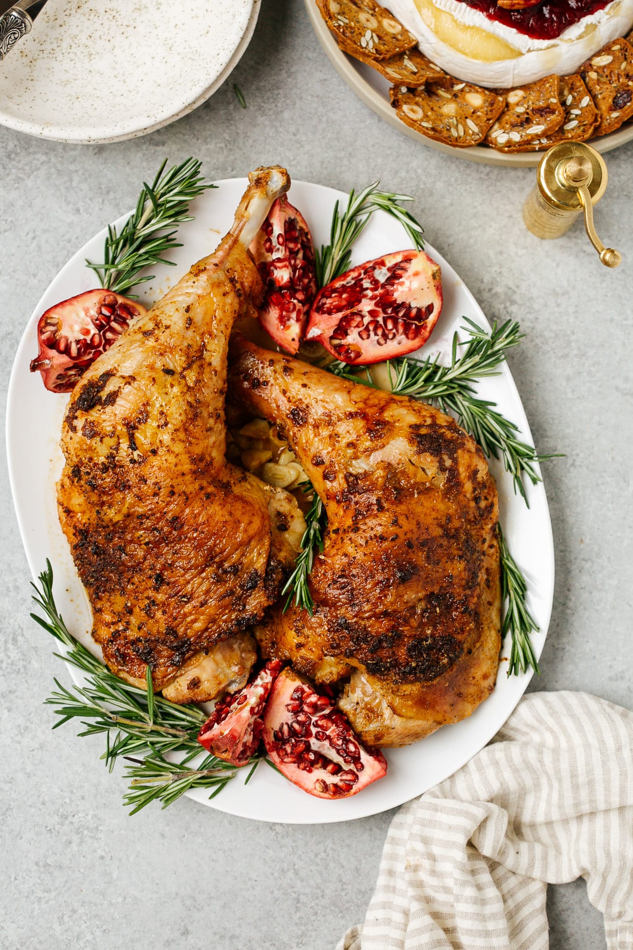 Overhead view of a platter with two roasted turkey legs surrounded by herbs and pomegranate. 