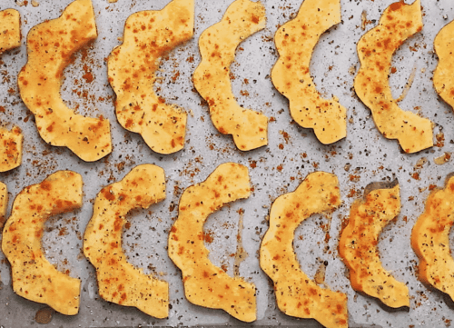 Sliced pieces of acorn squash, seasoned with spices, are arranged in rows on a baking sheet, ready for roasting.