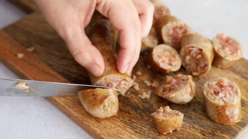 A hand uses a knife to slice cooked sausage into pieces on a wooden cutting board, preparing ingredients for Crockpot Sausage and Peppers.