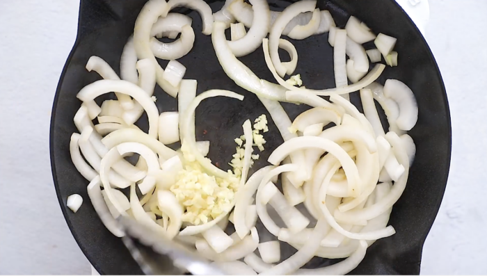 Sliced onions and minced garlic are being sautéed in a black skillet, the perfect base for Crockpot Sausage and Peppers, with tongs visible in the lower left corner.