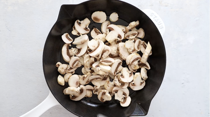 Sliced mushrooms being sautéed in a black cast iron skillet with a white handle, set on a light-colored surface—perfect for adding to your favorite Crockpot Sausage and Peppers recipe.