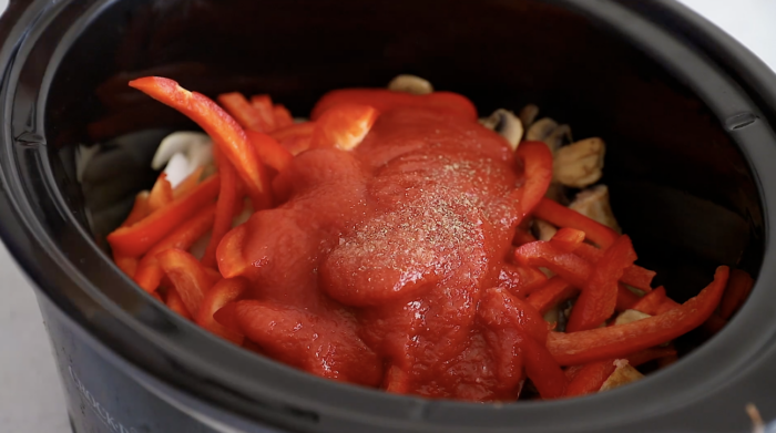 A slow cooker filled with sliced red bell peppers, mushrooms, tomato sauce, sausage, and seasonings—perfect for making hearty Crockpot Sausage and Peppers.