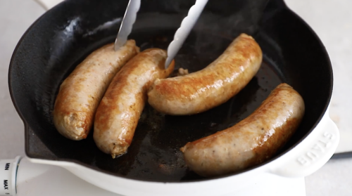 Four sausages are being cooked in a black skillet, with metal tongs turning one. The sizzling sausages are perfectly browned—a great start before making Crockpot Sausage and Peppers on your white stovetop.