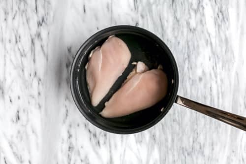 A black pot with two raw chicken breasts in water sits on a white marble surface, ready for boiling the perfect base for Sweet Potato Chicken Soup.
