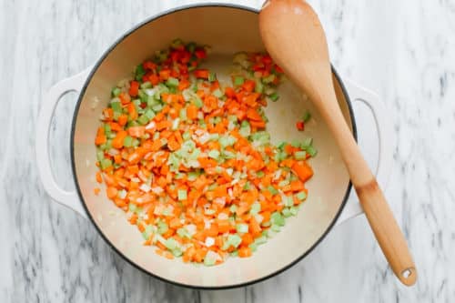 A white pot on marble holds chopped carrots, celery, and onions being cooked for Sweet Potato Chicken Soup, with a wooden spoon resting on the rim.