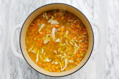 A white pot filled with Sweet Potato Chicken Soup, containing shredded chicken, diced carrots, tender sweet potatoes, and celery in a clear broth, sits on a marble surface.