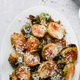 Oval white plate with Roasted Brussel Sprouts topped with grated Parmesan cheese. A small bowl of grated cheese sits in the corner. Text above reads Garlic Parmesan Brussels Sprouts.
