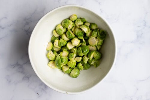 A white bowl filled with halved fresh Brussels sprouts, perfect for making Roasted Brussel Sprouts, sits on a white marble surface.