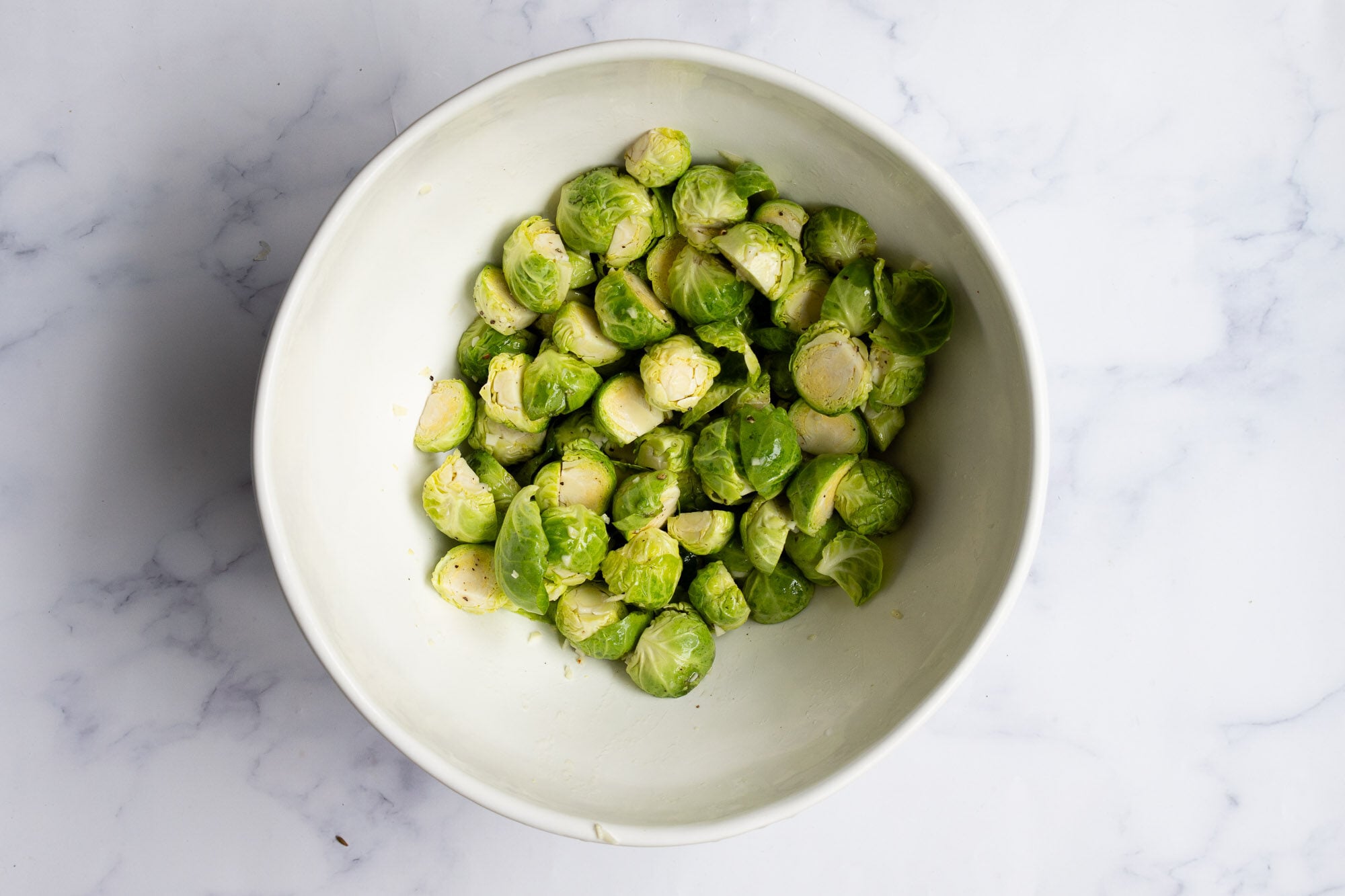 A white bowl filled with halved fresh Brussels sprouts, perfect for making Roasted Brussel Sprouts, sits on a white marble surface.