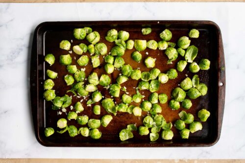 A baking sheet on a marble surface topped with halved Brussels sprouts and chopped garlic, ready to become perfectly roasted Brussels sprouts.