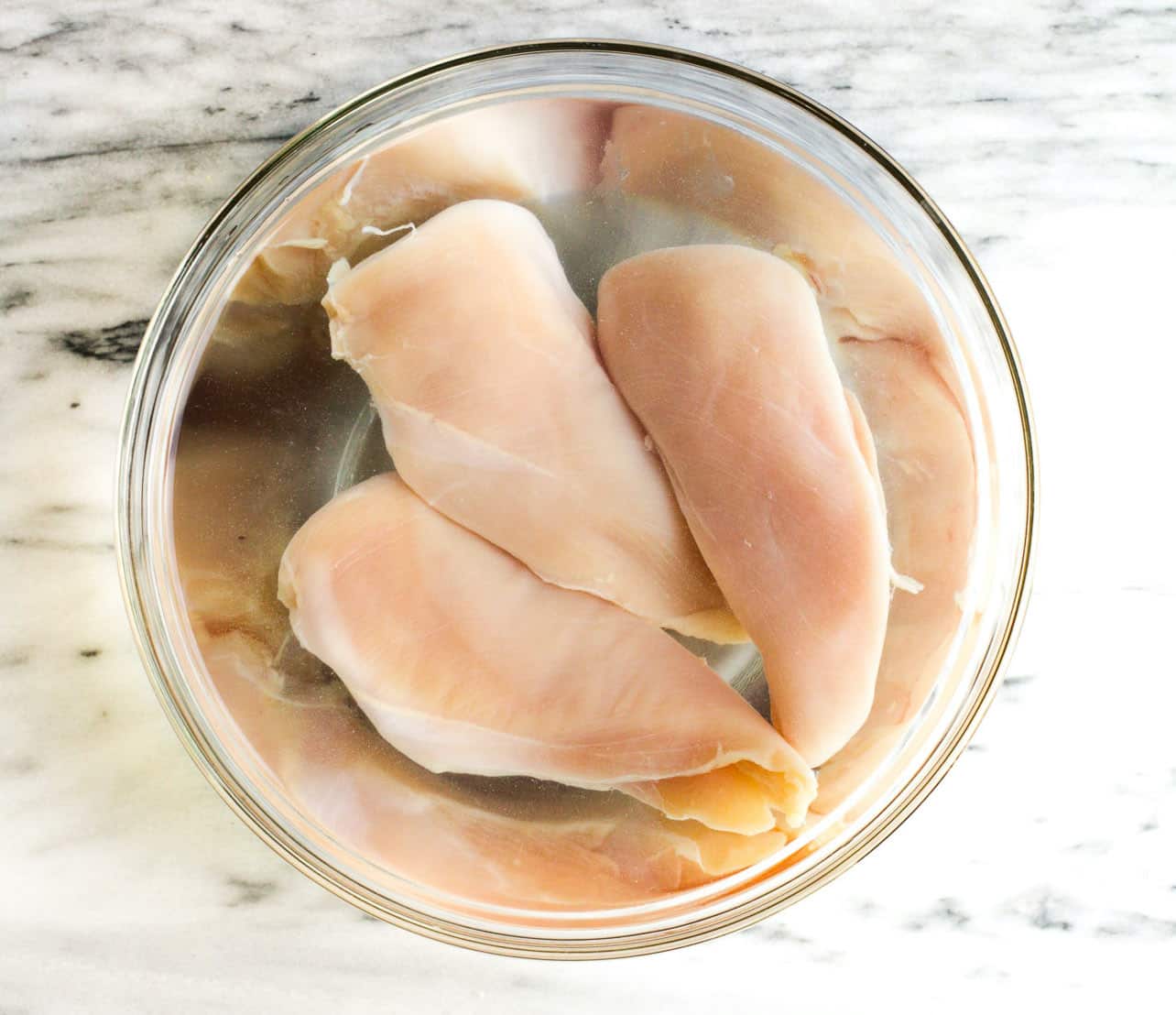 overhead view of raw chicken breast in a bowl with water and salt
