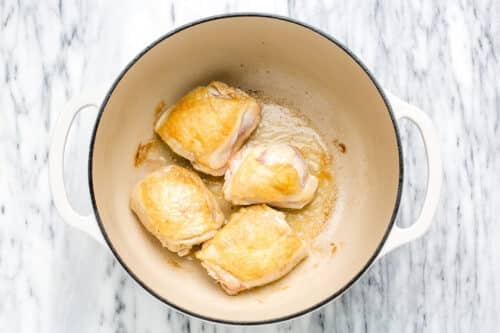 A white Dutch oven with three browned chicken thighs cooking inside, placed on a white marble surface—the perfect start for a comforting Chicken Noodle Soup.