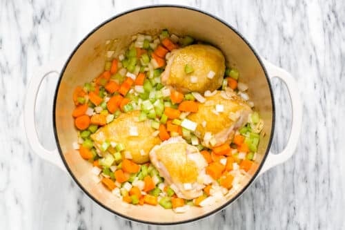 A white pot with browned chicken thighs, chopped carrots, celery, and onions on a marble surface—perfect for starting a comforting Chicken Noodle Soup.