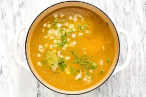 A pot of golden Chicken Noodle Soup topped with chopped onions, herbs, and a bay leaf sits on a white marble surface, seen from above.