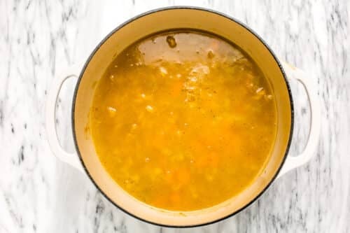 A large white pot filled with simmering yellow broth, brimming with small vegetable pieces and tender noodles, sits on a marble countertop—perfect for a comforting batch of chicken noodle soup.