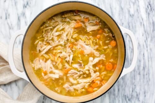 A large white pot filled with classic Chicken Noodle Soup, brimming with shredded chicken, carrots, and noodles, sits on a marble countertop with a beige cloth nearby.