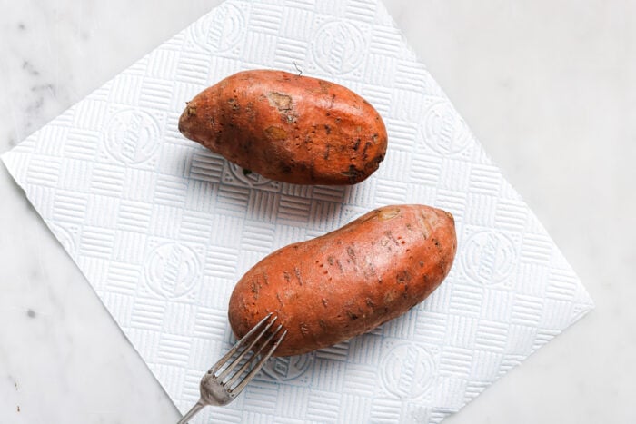 Two unpeeled sweet potatoes on a paper towel, with a fork next to one potato, rest on a white surface—perfect for making delicious stuffed sweet potatoes at home.