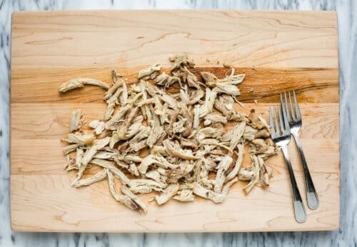 Shredded cooked chicken on a wooden cutting board with two forks beside it, on a marble countertop.