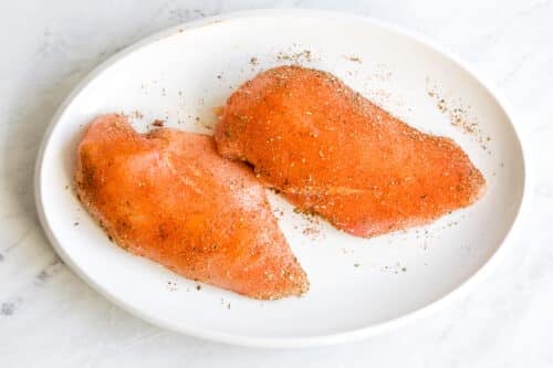 Two raw chicken breasts coated with seasoning, ready to become juicy and flavorful Instant Pot Chicken Breast, placed on a white round plate on a light marble surface.