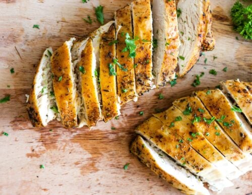 Two sliced, seasoned Instant Pot chicken breasts garnished with chopped parsley rest on a wooden cutting board, showcasing juicy, golden-brown edges and tender white meat.