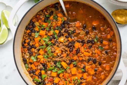 A pot of Sweet Potato Chili featuring ground meat, black beans, diced carrots, and fresh herbs, garnished with cilantro. Lime wedges are visible in the background.