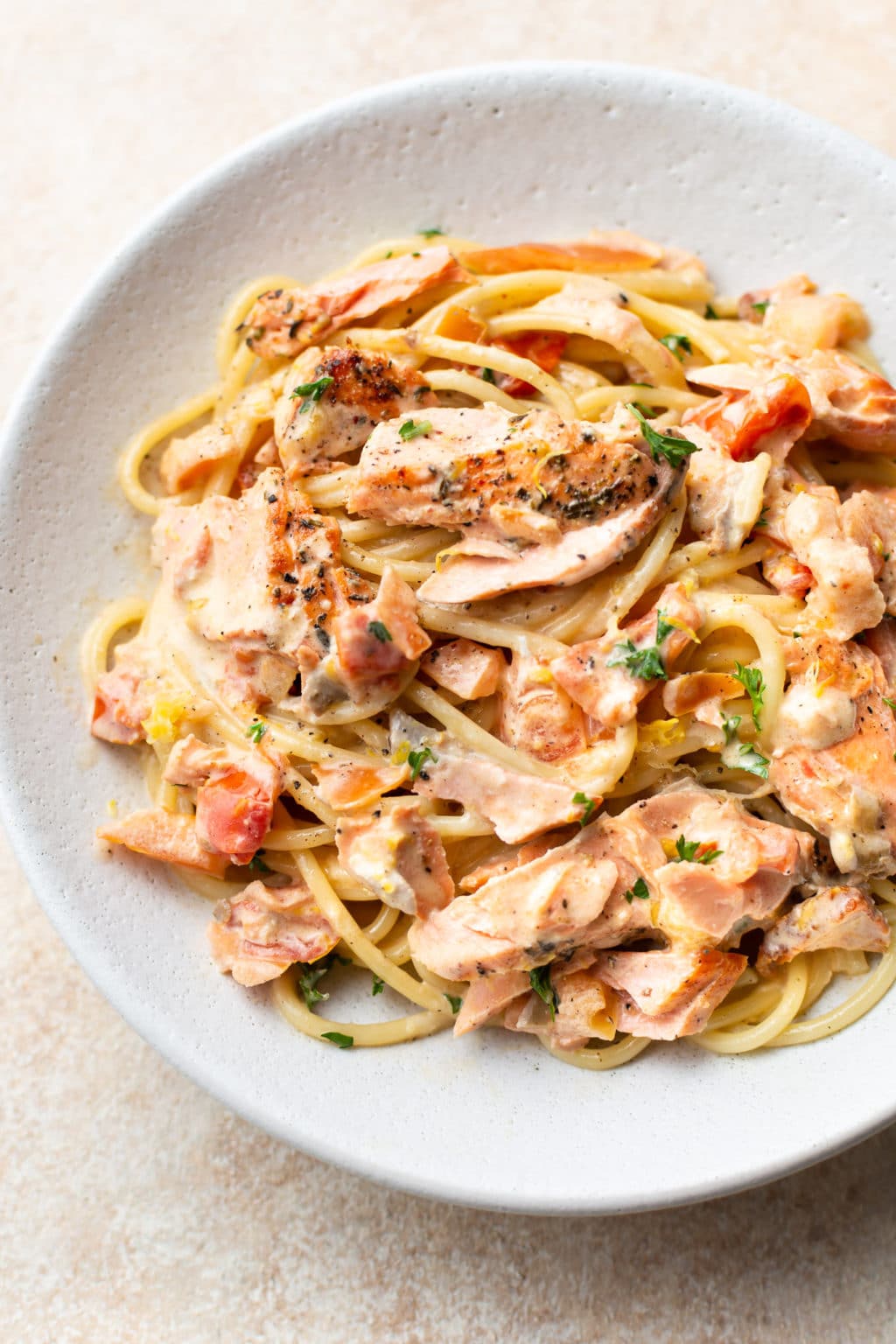 A white plate of spaghetti topped with creamy sauce, chunks of cooked salmon, diced tomatoes, and garnished with herbs—one of our favorite Easy Salmon recipes for a quick, delicious meal.