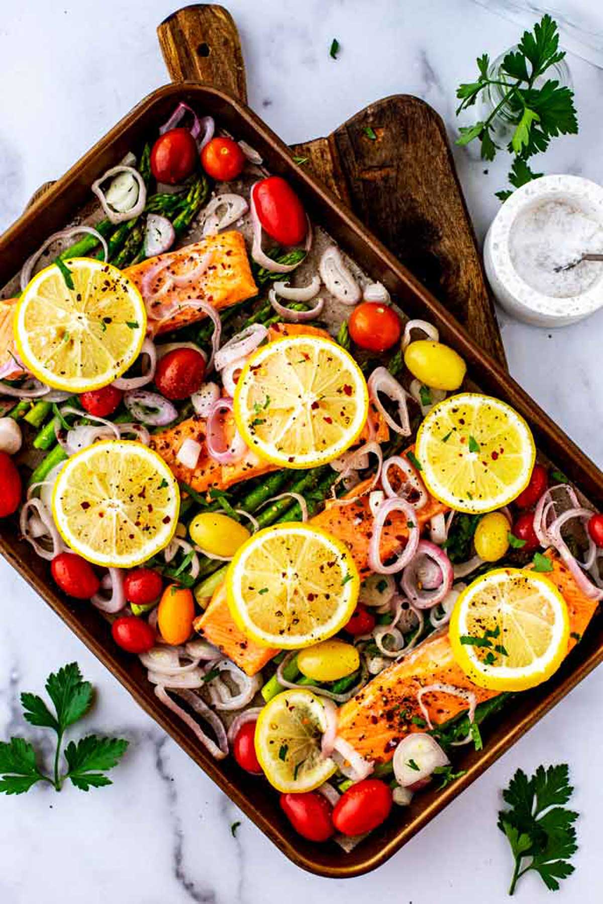 A baking tray with salmon fillets topped with lemon slices, surrounded by cherry tomatoes, asparagus, and sliced shallots—an inviting scene perfect for easy salmon recipes. Fresh herbs and a small bowl of salt rest nearby on the wooden board.