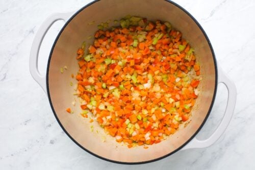 Diced carrots, celery, and onions are sautéing in a large, light-colored pot on a white marble countertop.