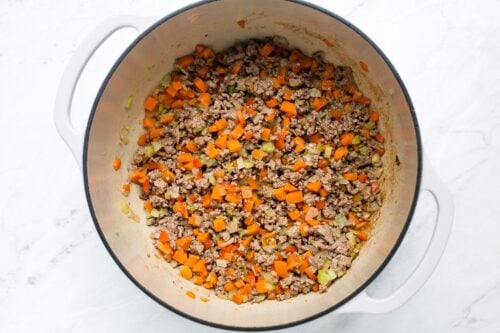 A large white pot filled with cooked ground meat, diced carrots, celery, and onions, sitting on a white marble surface. The ingredients are mixed evenly and lightly browned.