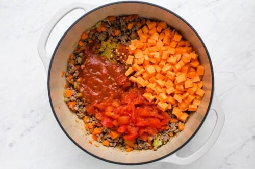 Large white pot on a marble surface filled with diced sweet potatoes, canned tomatoes, tomato sauce, cooked ground meat, chopped carrots, celery, onion, and spices, ready to be mixed for a stew or chili.