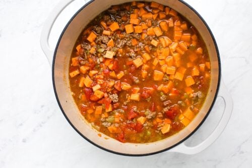 A white pot filled with a chunky stew containing diced sweet potatoes, ground meat, tomatoes, and vegetables, sitting on a white marble surface.