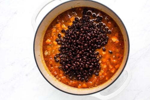 A white pot filled with a stew of diced vegetables and topped with black beans, sitting on a white marble surface.