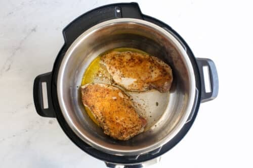 Overhead view of two seasoned chicken breasts cooking inside an Instant Pot on a white marble surface.