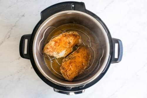 Two seasoned chicken breasts are placed on a metal rack inside an open Instant Pot, ready for cooking. The pot sits on a white marble countertop.