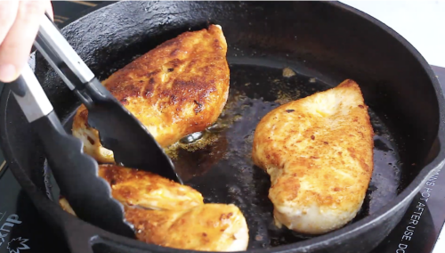 A close-up shot shows a hand using black tongs to flip golden-brown, seared chicken breasts in a hot, black cast-iron skillet on an induction stovetop, perfectly capturing the essence of this Mediterranean Chicken Skillet Recipe.